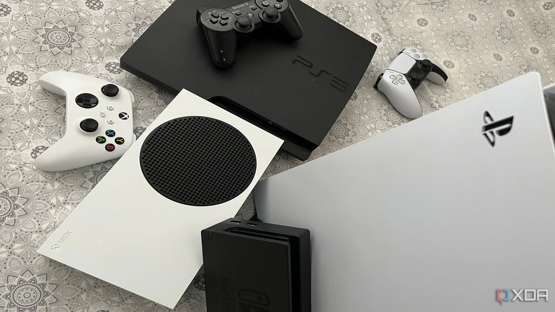 Bộ sưu tập các hệ máy console hiện tại và trước đây, bao gồm PS5, Xbox Series X, dock Switch và PS3, thể hiện sự đa dạng phần cứng trong ngành game.